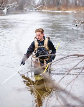 A person wades in a river, wearing a backpack and holding two poles, surrounded by bare trees.
